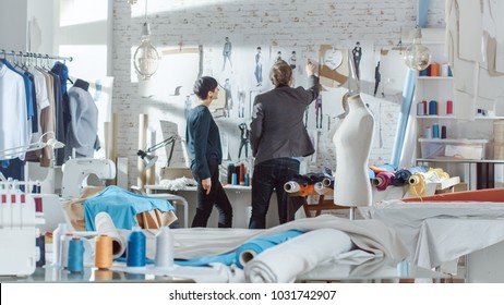 Female and Male fashion, designer,  Choosing and Pinning  to the Wall Clothing Sketches for Their New Collection. Studio is Sunny. Colorful Fabrics, Clothes Hanging and Sewing Items are Visible.