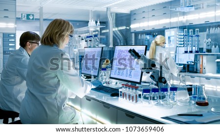 Female and Male Scientists Working on their Computers In Big Modern Laboratory. Various Shelves with Beakers, Chemicals and Different Technical Equipment is Visible.