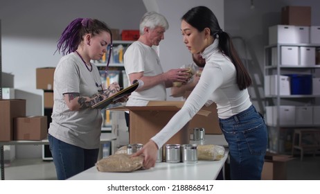 Female Manager Control Food Packing In Volunteer Center. Members Of Charity Organization Pack Food In Cardboard Boxes In Warehouse