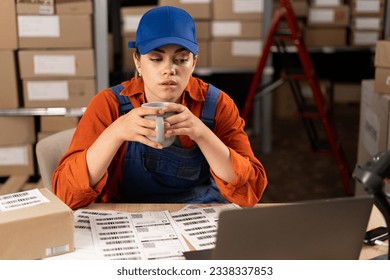 Female Manager drinking tea or coffee working in a warehouse storeroom with rows of shelves of parcels, packages ready for shipment. Break on work. Copy space - Powered by Shutterstock