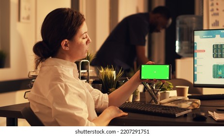 Female Manager Holding Smartphone With Greenscreen Display, Analyzing Blank Chroma Key Background On Mobile Phone. Using Isolated Mockup Template And Stock Market Exchange On Computer.