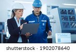 Female Manager and Project Engineer Wearing Hardhats Use Laptop in Industrial Factory, Talk, Plan Productivity Optimization. Production Line Workers Operate CNC Machinery, Program robot arm