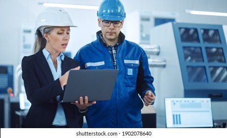 Female Manager And Project Engineer Wearing Hardhats Use Laptop In Industrial Factory, Talk, Plan Productivity Optimization. Production Line Workers Operate CNC Machinery, Program Robot Arm