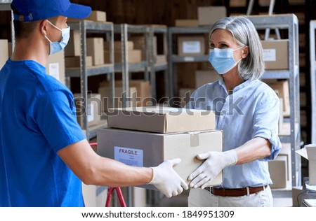Female manager supervisor wearing face mask preparing fast drop shipping safe delivery giving parcels packages boxes to male courier taking ecommerce orders to deliver standing in warehouse storage.