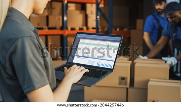Female Manager Using Laptop Computer To Check\
Inventory. In the Background Warehouse Retail Center with Cardboard\
boxes, e-Commerce Online Orders, Food, Medicine, Products Supply.\
Over the Shoulder