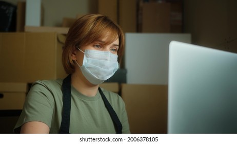 Female Manager Of Warehouse In Protective Medical Mask Working With Laptop, Making Business Correspondence With Clients, Sending Orders. Small Delivery Company Concept.