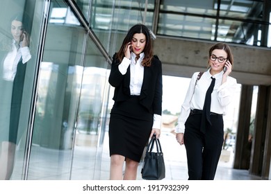 Female Managers Meet In A Business Context Outside Near A Modern Building Made Of Mirrors: They Both Talk On The Phone While Walking