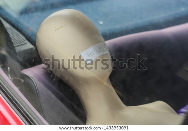 Female mannequin with no eyes,\
nose or mouth in a car with \'help\' sign where should be the\
lips