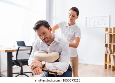 Female Massage Therapist Massaging Back Of Businessman Sitting On Massage Chair In Office On Blurred Background