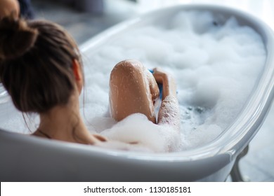 female massaging her legs with sponge in the tub. back view shot