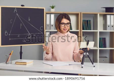 Female math and geometry teacher explains new material to students through a video call using a phone on a tripod. Woman sits in a classroom with a blackboard next to her and conducts an online lesson