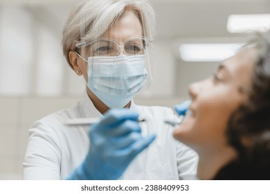 Female mature stomatologist dentist orthodont curing healing boy`s teeth tooth, doing mouth cavity cleaning, removing baby tooth - Powered by Shutterstock