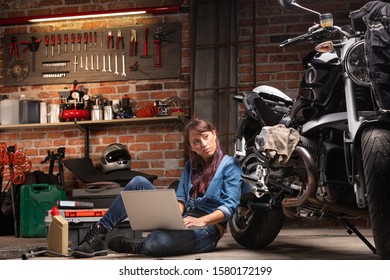 Female Mechanic Checking For Vintage Motorbike Parts On A Laptop Computer As She Services Or Repairs A Bike In A Spacious Workshop