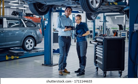 Female Mechanic Talking to a Manager Under a Vehicle in a Car Service. Specialist is Showing Info on a Tablet Computer. Empowering Woman Wearing Gloves and Safety Gloves. Modern Clean Workshop. - Powered by Shutterstock