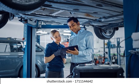 Female Mechanic Talking to a Manager Under a Vehicle in a Car Service. Specialist is Showing Info on a Tablet Computer. Empowering Woman Wearing Gloves and Safety Gloves. Modern Clean Workshop. - Powered by Shutterstock