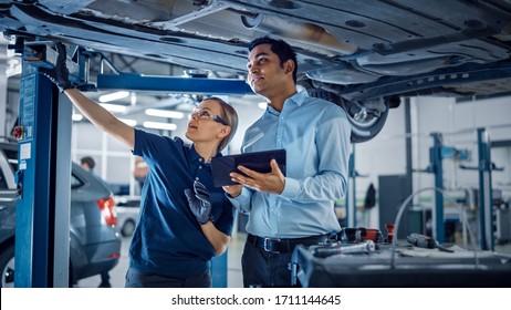 Female Mechanic Talking to a Manager Under a Vehicle in a Car Service. Specialist is Showing Info on a Tablet Computer. Empowering Woman Wearing Gloves and Safety Gloves. Modern Clean Workshop.