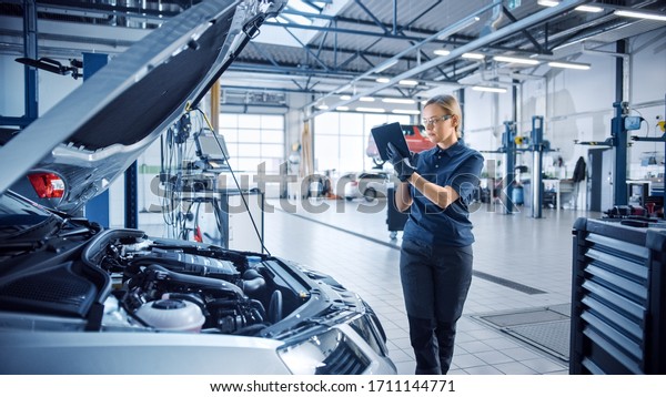 Female Mechanic Uses a Tablet Computer with an
Augmented Reality Diagnostics Software. Specialist Inspecting the
Car in Order to Find Broken Components Inside the Engine Bay.
Modern Car Service.