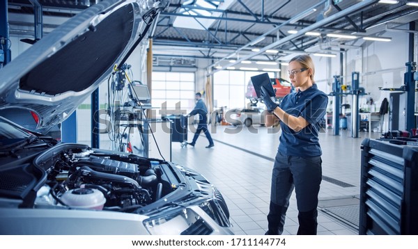 Female Mechanic Uses a Tablet Computer with an\
Augmented Reality Diagnostics Software. Specialist Inspecting the\
Car in Order to Find Broken Components Inside the Engine Bay.\
Modern Car Service.