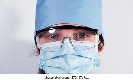 Female Medic In Disposable Medical Mask, Protective Plastic Glasses And Textile Cap, Portrait Of A Tired Doctor With Sad Eyes, Close Up