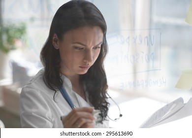 Female Medical Doctor Working At Clinic Office. Writing On Glass Whiteboard Symptoms And Test Results Of Her Patient To Diagnose Disease