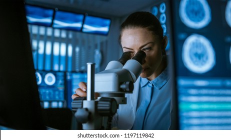 Female Medical Research Scientist Looking Through the Microscope Types Acquired Data in the Computer. Laboratory. In the Laboratory with Multiple Screens Showing MRI / CT Brain Scan Images.