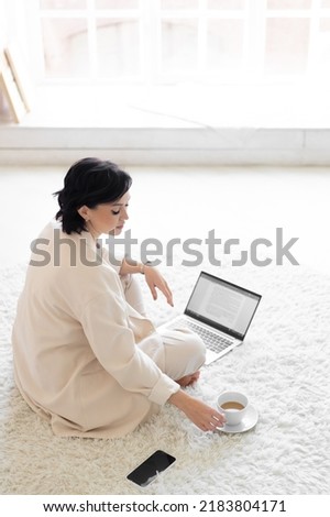 Female milf with black hair in a cream suit sits on a cozy carpet on the floor. Coffee break. Working on a laptop in a minimalist home living room.