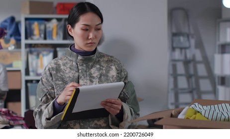 Female Military Officer With Clipboard Supervise Work Of Volunteers In Warehouse. Diverse People Sort And Pack Donated Goods For Humanitarian Aid