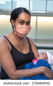 A female mixed race African American mother in a black tank top wears a makeshift striped orange fabric mask and holds her infant baby girl wrapped in her blue jacket during the COVID-19 pandemic.