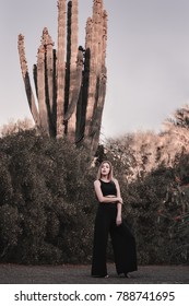 Female Model With Blond Hair And In Black Trendy Suit Standing Near A Very Tall Cactus. Hippie And Boho Style