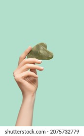 Female Model Hand With Green Jade Gua Sha Scraper On Green Background. Spa Body Care Tool. Beauty Care Gua Sha Stone Scraper. Copy Space