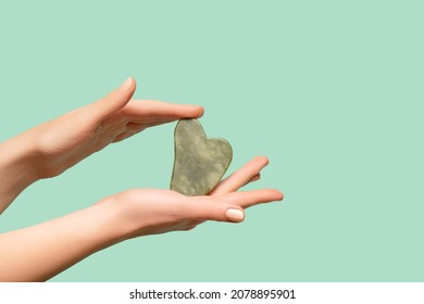 Female Model Hands With Green Jade Gua Sha Scraper On Green Background. Spa Body Care Tool. Beauty Care Gua Sha Stone Scraper. Copy Space