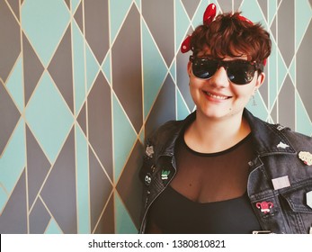 A Female Model In Her 20's Wearing A Black Denim Jacket Covered In Enamel Pins. She Is Also Wearing A Red Polka Dot Bow And Sunglasses. The Shot Was Taken Against A Patterned Wallpaper.