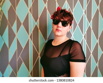 A Female Model In Her 20's Wearing A Black Denim Jacket Covered In Enamel Pins. She Is Also Wearing A Red Polka Dot Bow And Sunglasses. The Shot Was Taken Against A Patterned Wallpaper.