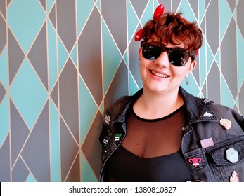 A Female Model In Her 20's Wearing A Black Denim Jacket Covered In Enamel Pins. She Is Also Wearing A Red Polka Dot Bow And Sunglasses. The Shot Was Taken Against A Patterned Wallpaper.