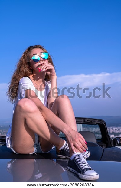 Female model posing and having fun\
siting on back of a convertible car at sunny summer\
day.