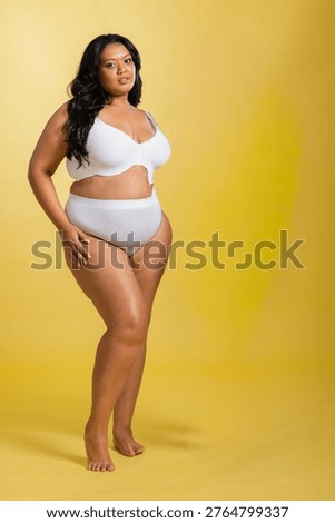 Female model standing and posing on yellow backdrop and floor wearing white bra high-waist briefs. Minimalism, serenity, studio, softlight, lingerie, barefoot, centered