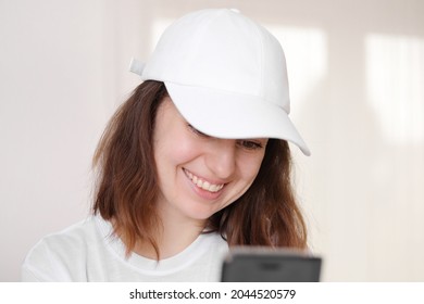 Female Model Wearing A White Baseball Cap. White Cap Mockup, Template For Picture, Text Or Logo. Girl With A Cap Holding Smarthphone. Free Space, Copy Space.