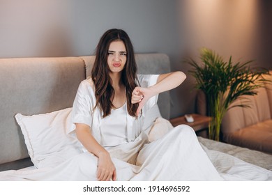 Female Model In White Silk Pajamas Showing Dislike. Beautiful Woman With Sad Face Sitting On The Bed Shows Dislike