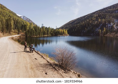 Female Motorcyclist With Her Motorcycle Standing On The Road Near Mountain Lake, Clear Reflection On Blue Water, Autumn Colors Of Trees, Sunny Day