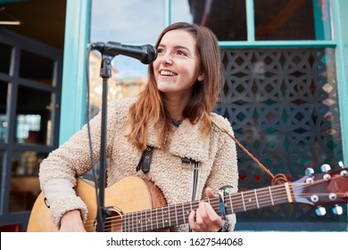 Female Musician Busking Playing Acoustic Guitar And Singing Outdoors In Street