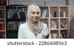 Female muslim student in library, holding books and looking at camera, girl wearing hijab spending time studying - modern Islam, student lifestyle concept close up 