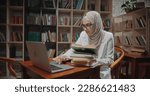 Female muslim student is studying at desk in library, using laptop and books. Girl wearing hijab is preparing for exams - student lifestyle, modern Islam concept 4k close 
