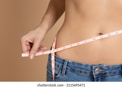 Female naked belly with tape in hands measuring waistline with blue jeans on beige background. Keeping diet and exercising. Setting and achieving goals. 