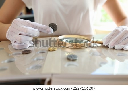 Female numismatist examines collection of coins. Woman looks at coins through magnifying glass. Numismatic collection review.
