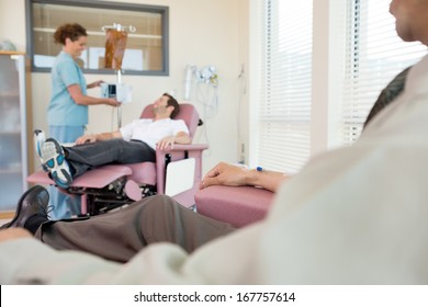 Female Nurse Adjusting IV Machine For Patient Reclining On Chair In Hospital Room