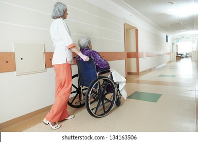 Female Nurse Carer And Aged Elderly Patient Woman In Wheelchair At Clinic Hallway