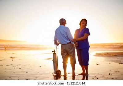 Female Nurse Comforting A Senior Male Patient Standing With His Oxygen Tank On A Beach At Sunset.