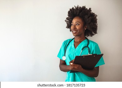 Female Nurse Or Doctor Smiles While Staring Out Window In Hospital Hallway And Holding Clipboard With Patient File. African American Female Pediatric Nurse In Office. Portrait Of Female Nurse Standing