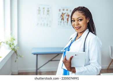 Female Nurse Or Doctor Smiles While Staring Out Window In Hospital Hallway And Holding Digital Tablet With Electronic Patient File. Portrait Of Female African American Doctor Standing In Her Office 