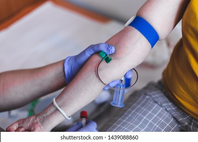 Female Nurse Drawing Blood In A Plastic Vial From A Young Person With Blue Tourniquet In Hospital – Woman Doctor Taking Fluid Samples With Small Needle From Patient Hand